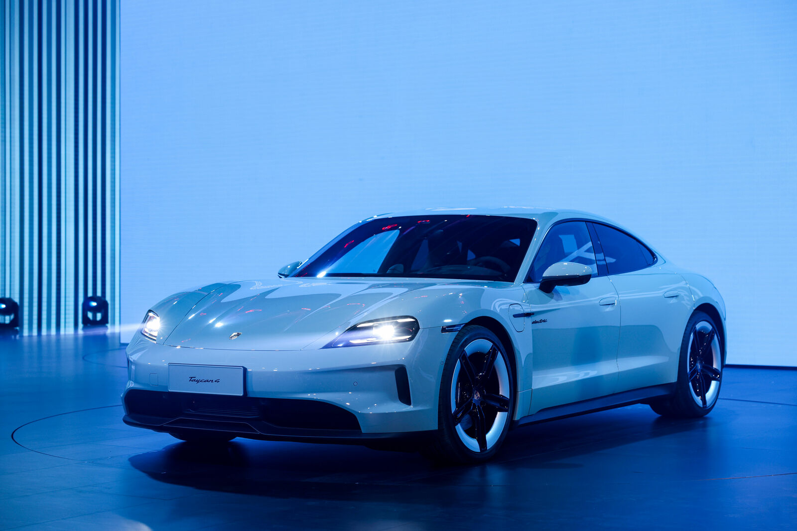 A sleek, white Porsche Taycan 4S is displayed in a brightly lit showroom with a futuristic blue background.