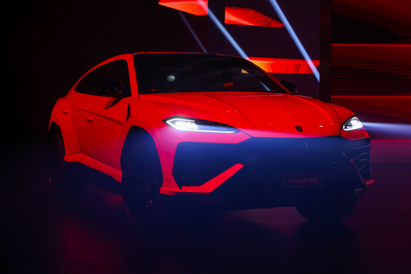 Ein knallroter Lamborghini Urus S SUV wird dramatisch auf der Bühne beleuchtet, mit schwarzem Hintergrund und betont durch leuchtend rote Beleuchtung.