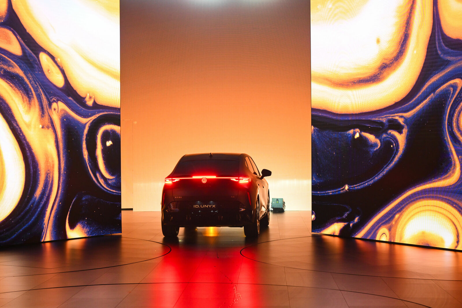 An image of a modern car displayed in a showroom against a backdrop of glowing, abstract artworks. The rear view of the car is illuminated, highlighting the red tail light prominently.