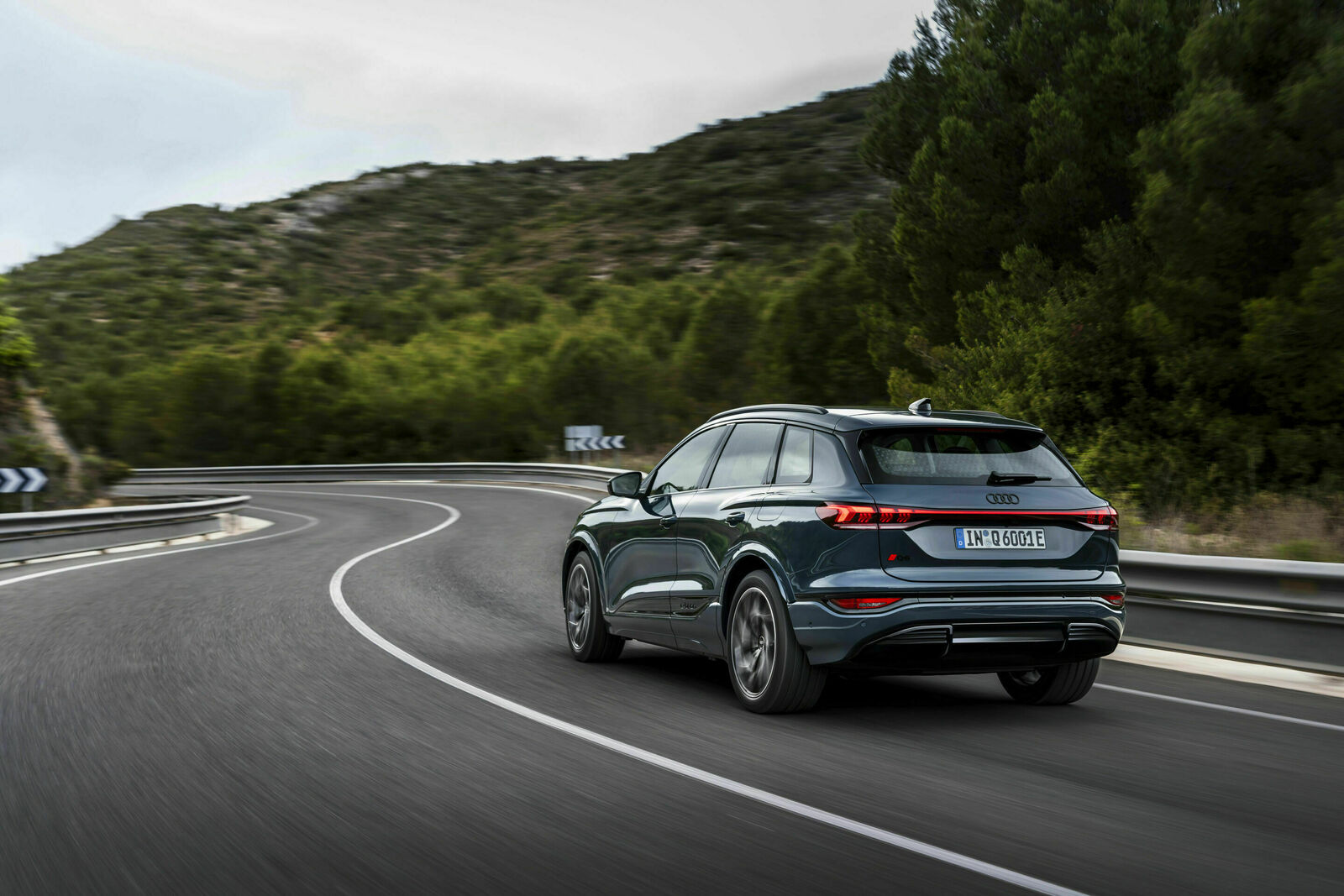 An image of a dark gray Audi Q6 e-tron speeding on a curvy mountain road. The environment is green with trees and shrubs along the road.