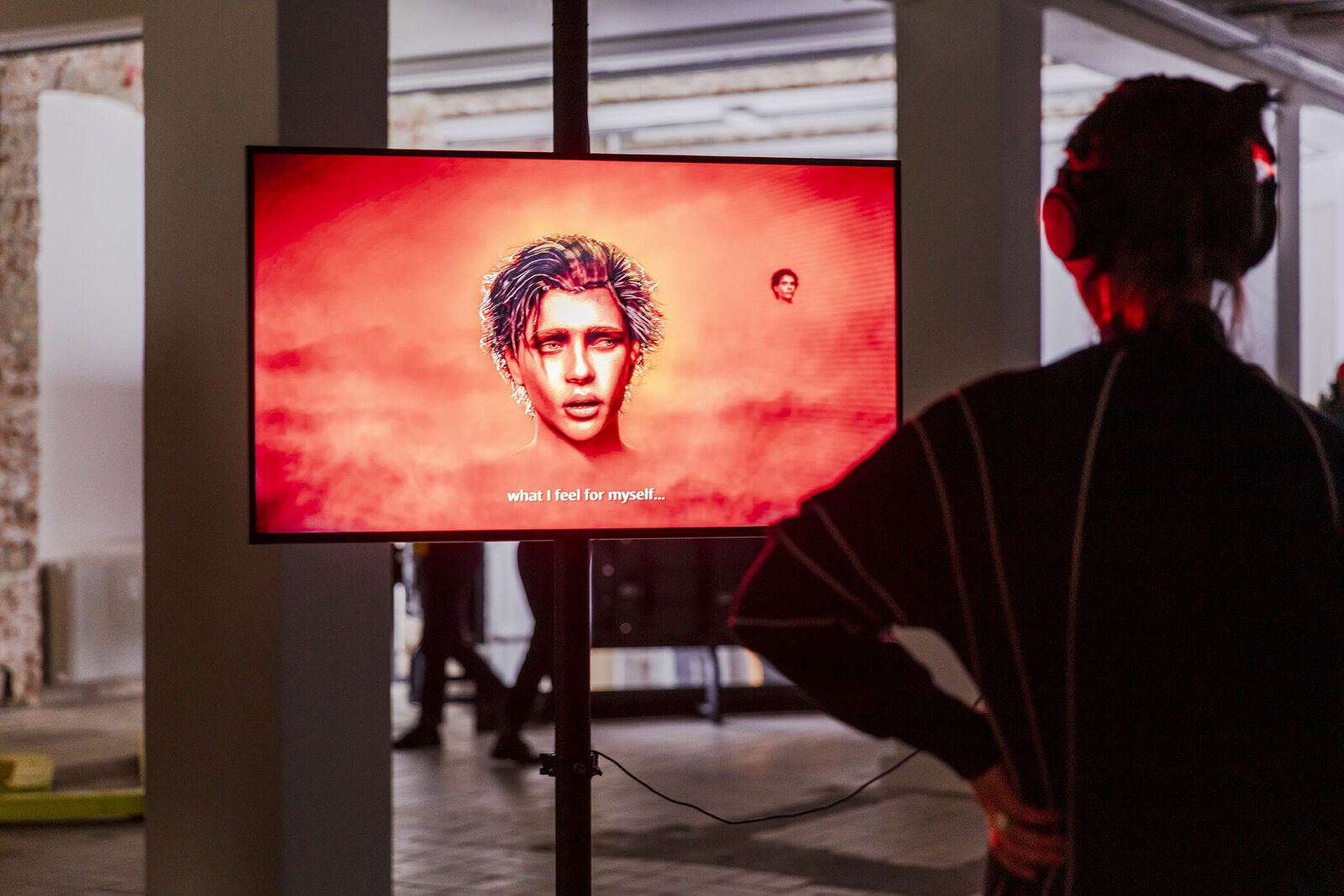 Eine Person steht in einer Ausstellung und betrachtet ein digitales Kunstwerk auf einem Bildschirm, das ein Gesicht vor einem roten Hintergrund zeigt, begleitet von dem Text "what I feel for myself...".