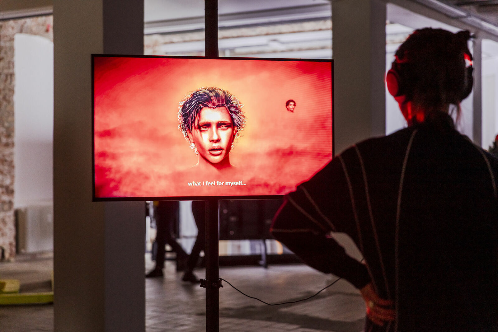 A person stands in an exhibition and looks at a digital artwork on a screen that shows a face in front of a red background, accompanied by the text "what I feel for myself...".