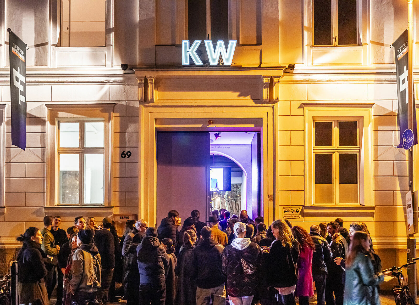 Menschenmenge vor dem Eingang eines Gebäudes mit der Leuchtreklame "KW" bei Nacht.