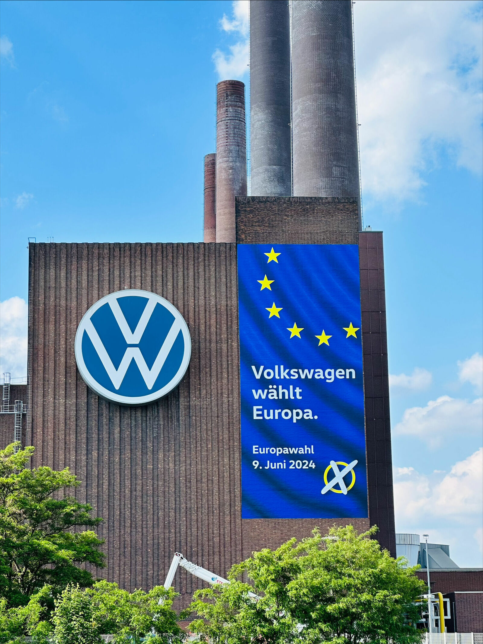 Ein großes Volkswagen-Werksgebäude mit einem Banner, das für die Europawahl am 9. Juni 2024 wirbt, mit einem blauen Hintergrund und gelben Sternen der EU-Flagge.