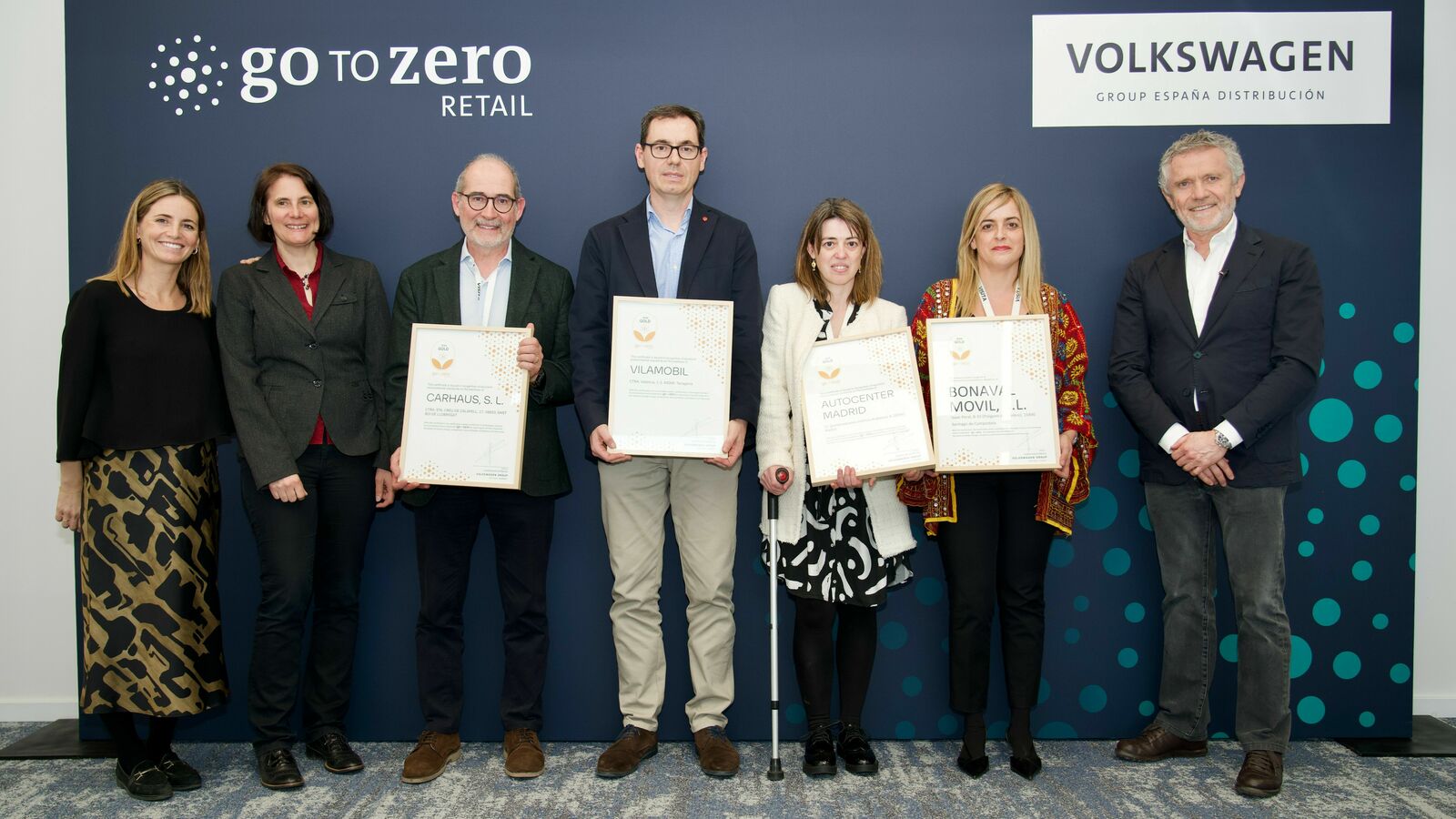 Group of six individuals proudly displaying certificates in front of a 'Go to Zero Retail' backdrop of the Volkswagen Group.