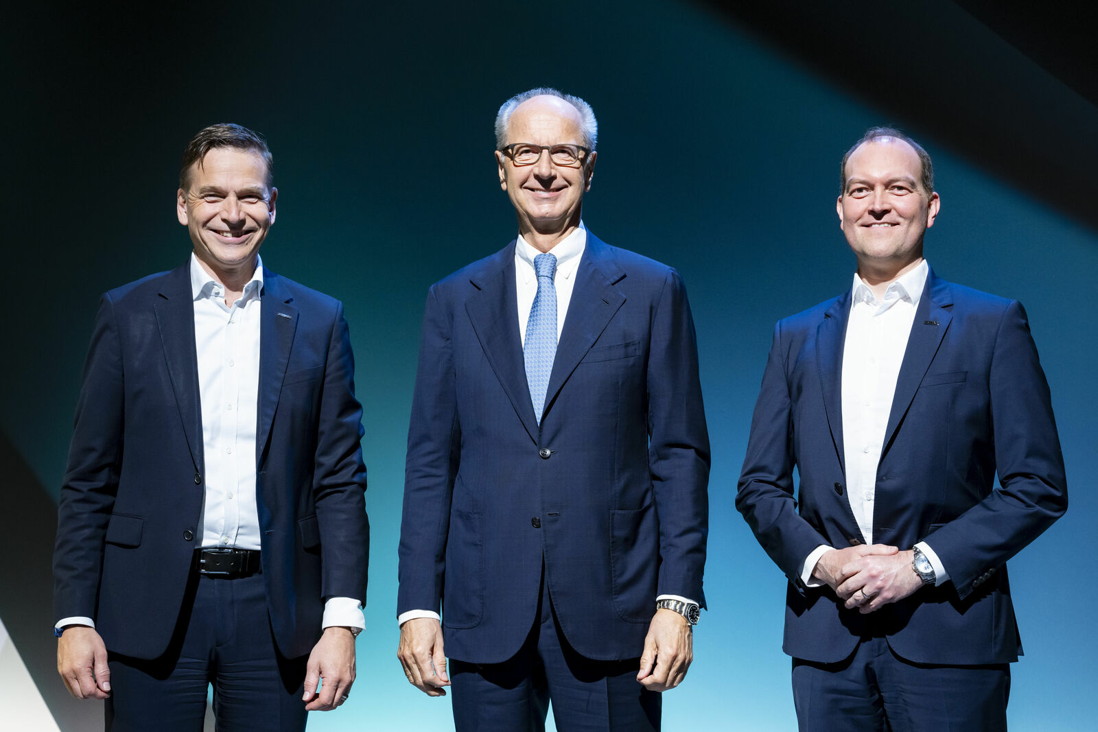 Three men in suits smiling and standing side by side on a stage with a dark background.