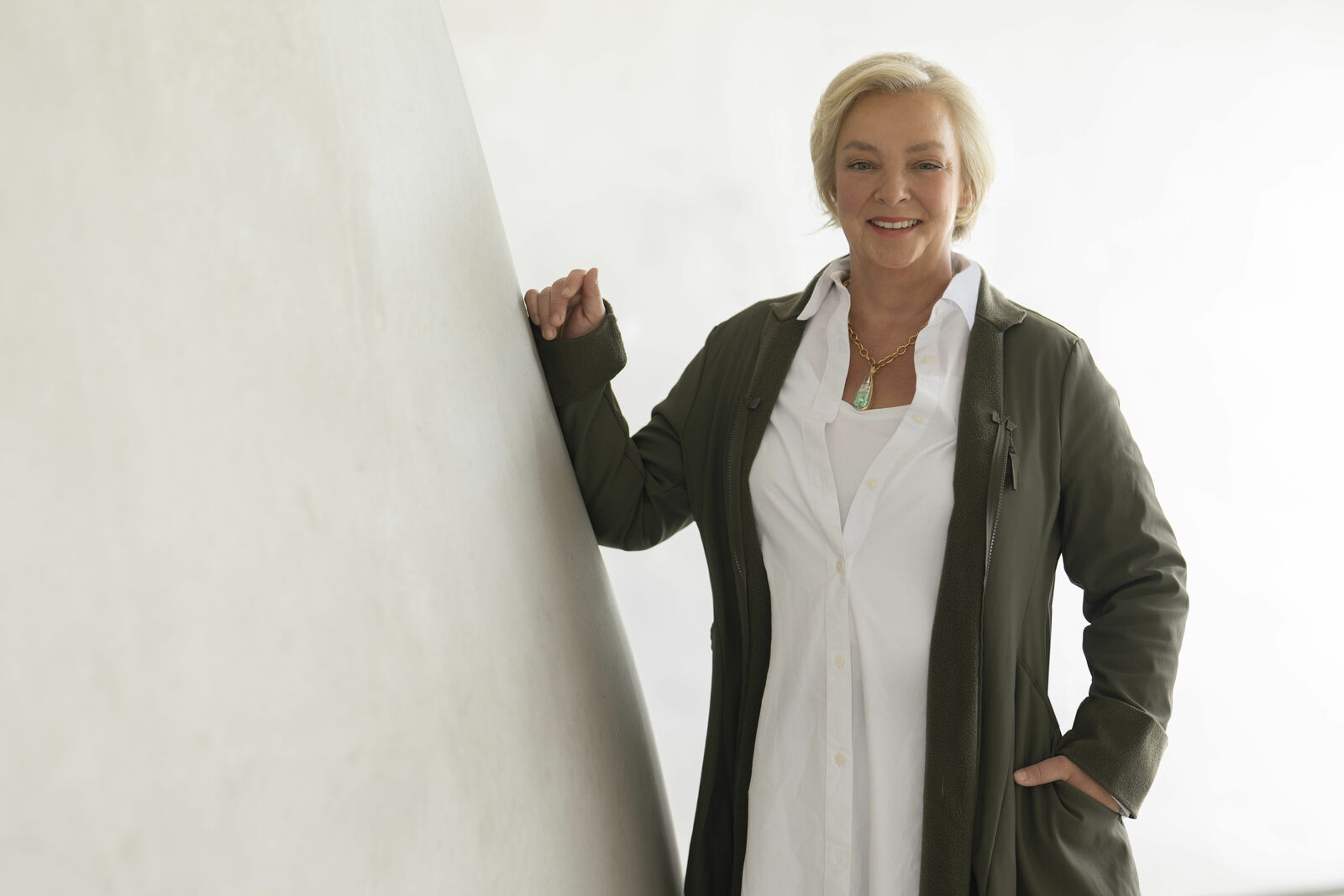 Porträt einer Frau mit blonden Haaren, die eine grüne Jacke und eine weiße Bluse trägt und lächelnd neben einer weißen Säule steht.