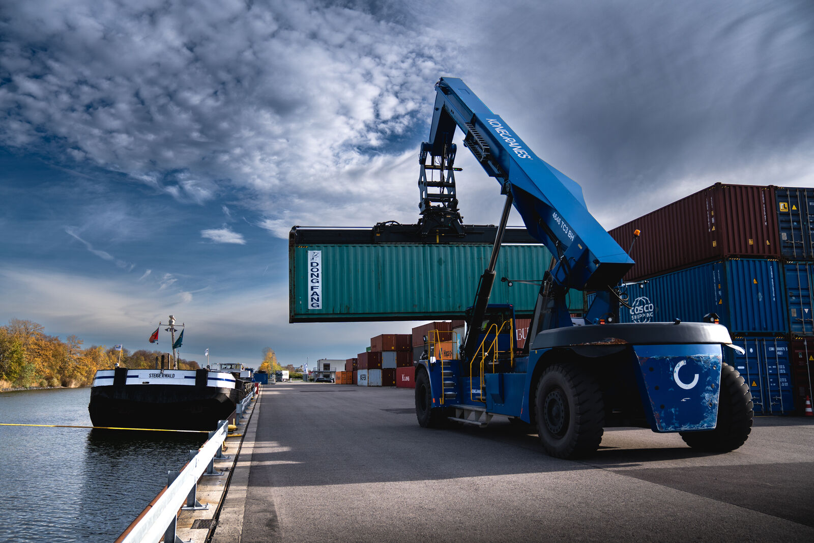 A large blue reach stacker lifts a green shipping container labeled "VOLKSWAGEN" at a container terminal. In the background, a cargo ship is docked along the quay, with stacked containers and a partly cloudy sky visible. The setting suggests a port or logistics hub where goods are transferred between water and land transportation.