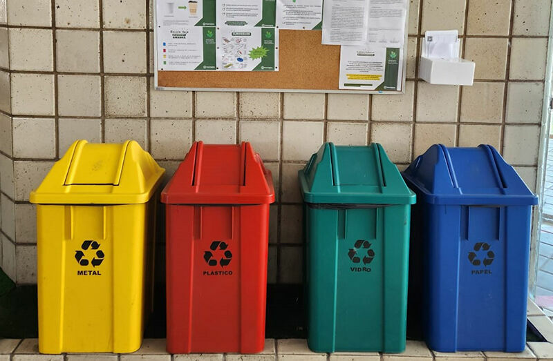 Four colorful recycling bins stand side by side: yellow for metal, red for plastic, green for glass, and blue for paper. A tiled wall with a bulletin board is visible in the background.