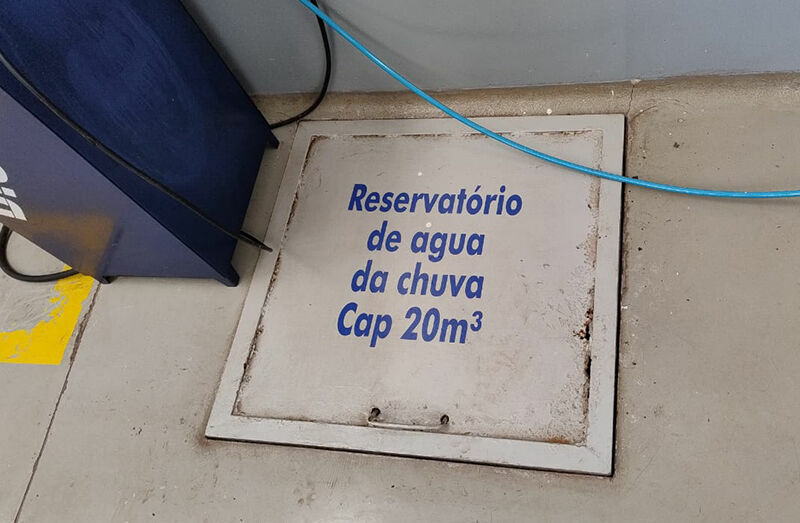 A floor hatch with the inscription "Reservatório de água da chuva Cap 20m³," indicating a rainwater reservoir with a capacity of 20 cubic meters. In the background, a blue container and a blue cable are visible.
