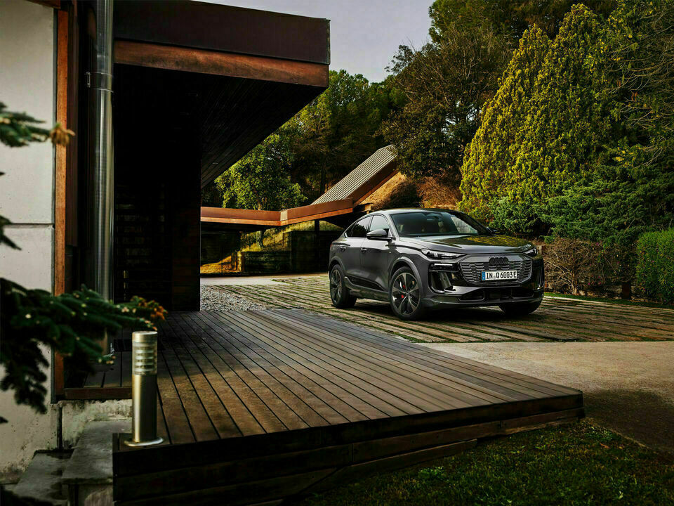 Ein schwarzer Audi SUV steht vor einem modernen Gebäude in einer natürlichen, grünen Umgebung. Das Fahrzeug ist auf einem Holzdeck geparkt, das an einen gepflegten Garten grenzt.