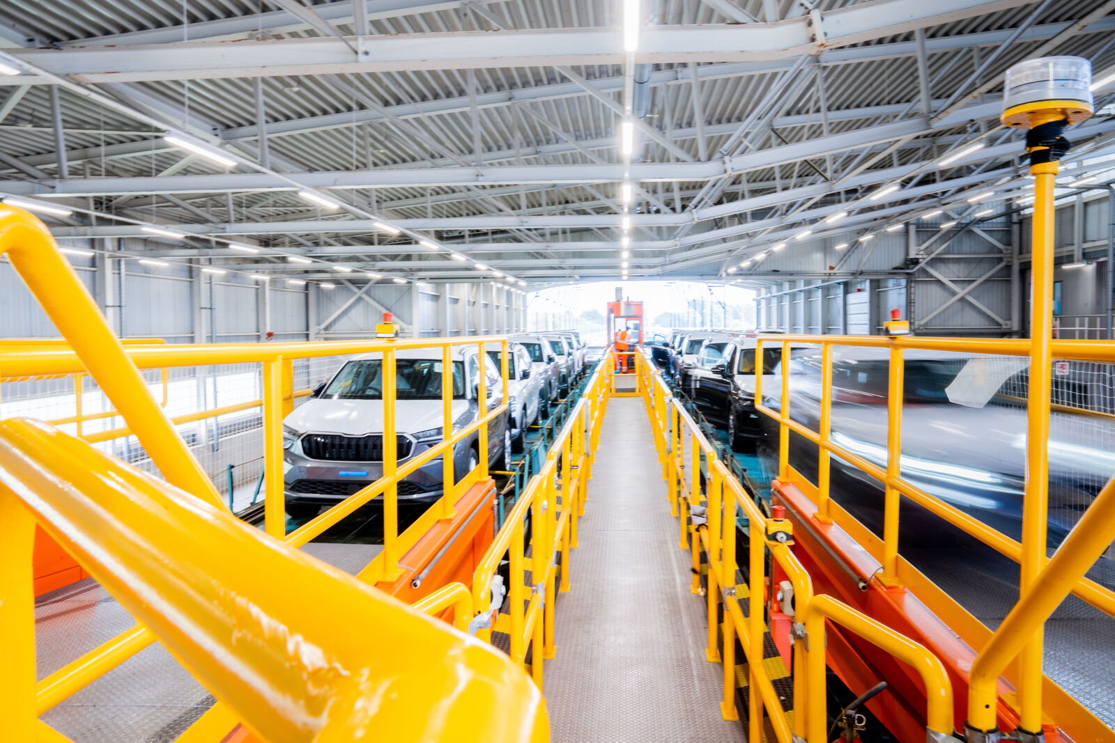 View through an industrial vehicle loading station with yellow safety railings. Several new cars, including white and black SUVs, are positioned on a conveyor belt or platform for loading. In the background, an open hall with daylight is visible, while some vehicles appear to be in motion, indicated by motion blur.