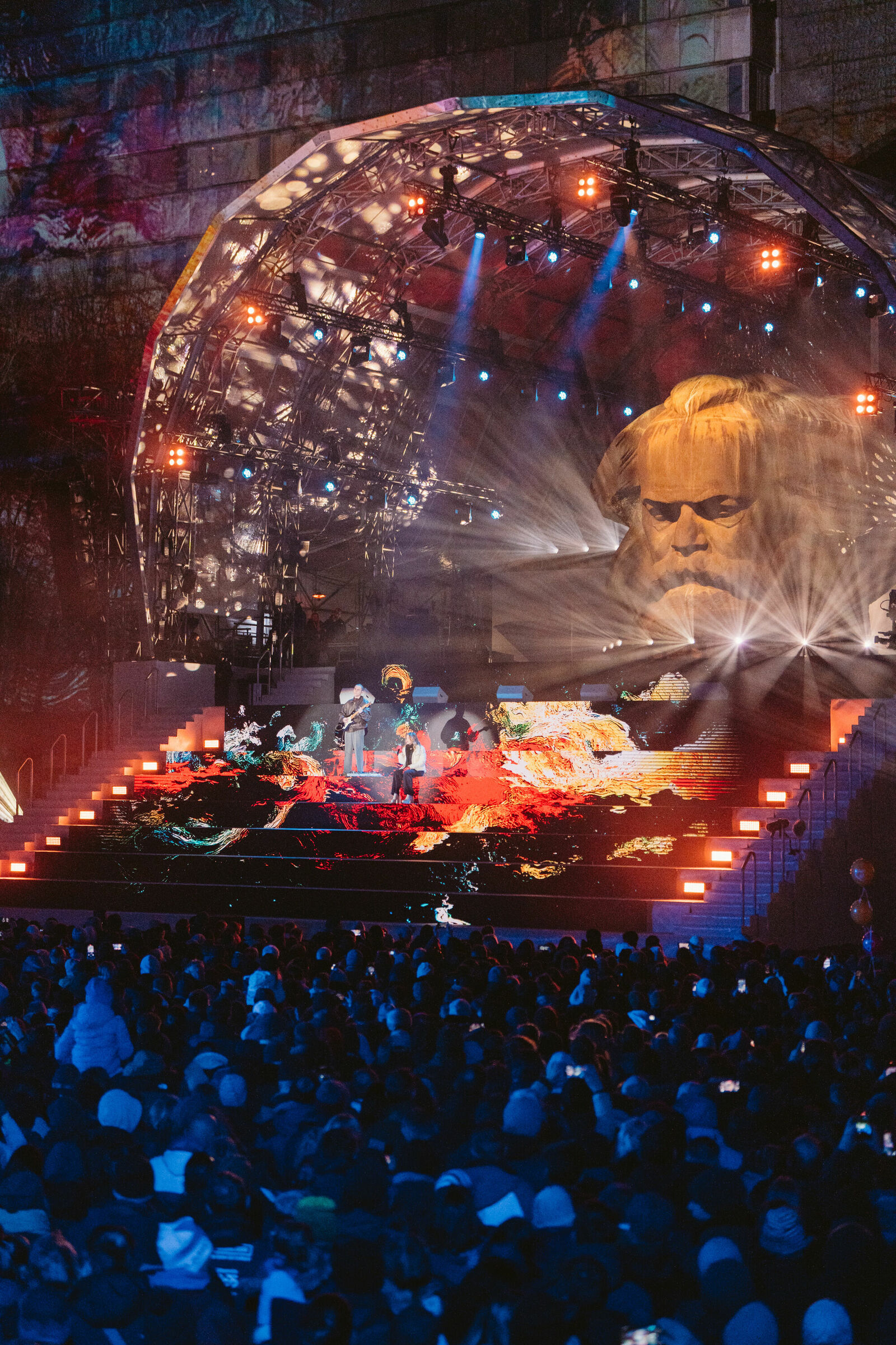 Eine große Bühne bei einer nächtlichen Veranstaltung, beleuchtet mit bunten Lichtern und einer Projektion eines Porträts auf der Bühne, von Karl Marx. Künstler performen vor einem Publikum, während abstrakte farbige Muster auf der Bühne projiziert werden. Die Veranstaltung findet unter einer beeindruckenden Kuppelkonstruktion statt.