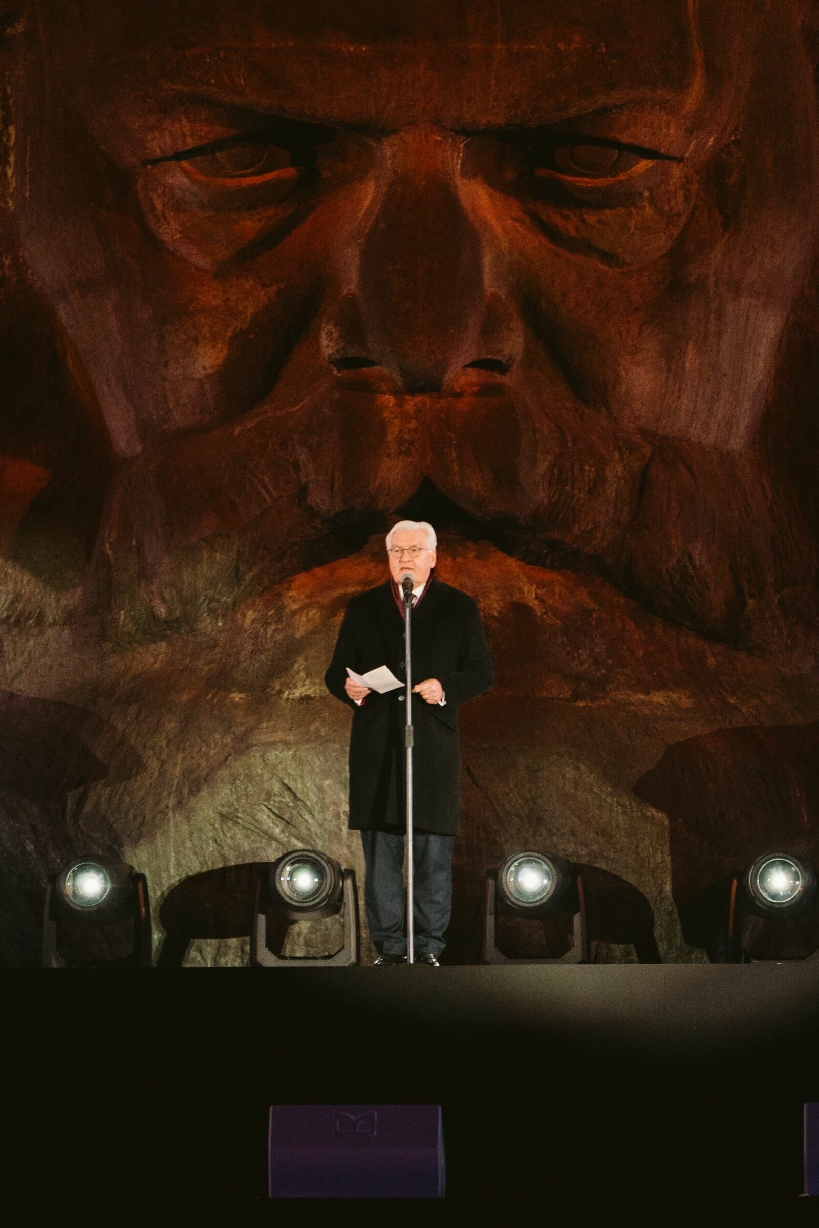 A man in a black coat is giving a speech at a microphone, holding a sheet of paper in his hand. In the background, a large stone-carved sculpture of a bearded face is dramatically illuminated. Four floor spotlights light up the scene.