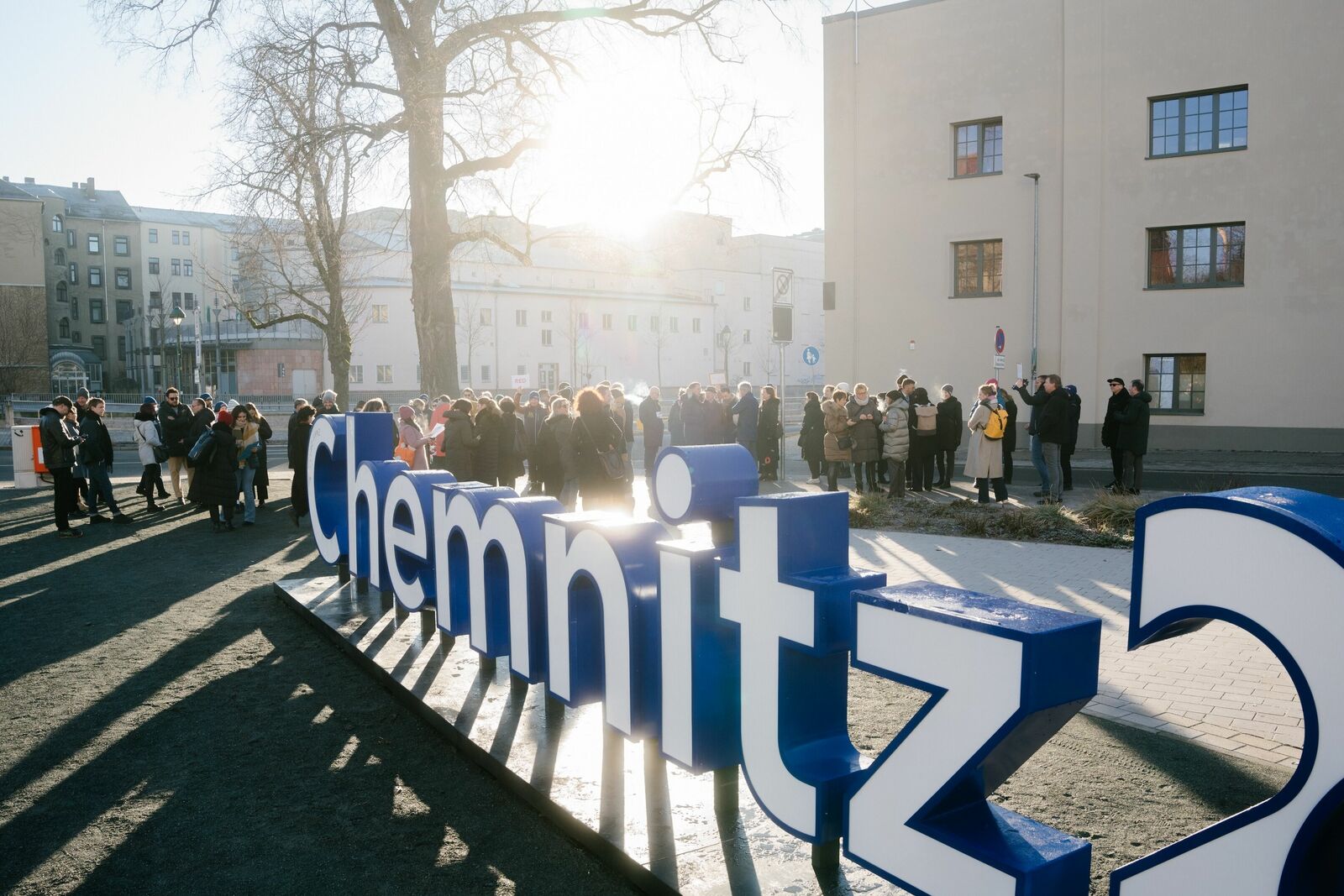 Das Bild zeigt eine Gruppe von Menschen, die sich vor einem großen, blauen "Chemnitz"-Schild versammelt haben. Die Sonne scheint von hinten, wodurch die Szenerie warm beleuchtet wird. Die Leute tragen Winterkleidung und stehen in einer Art Warteschlange, während ein Gebäude im Hintergrund zu sehen ist.