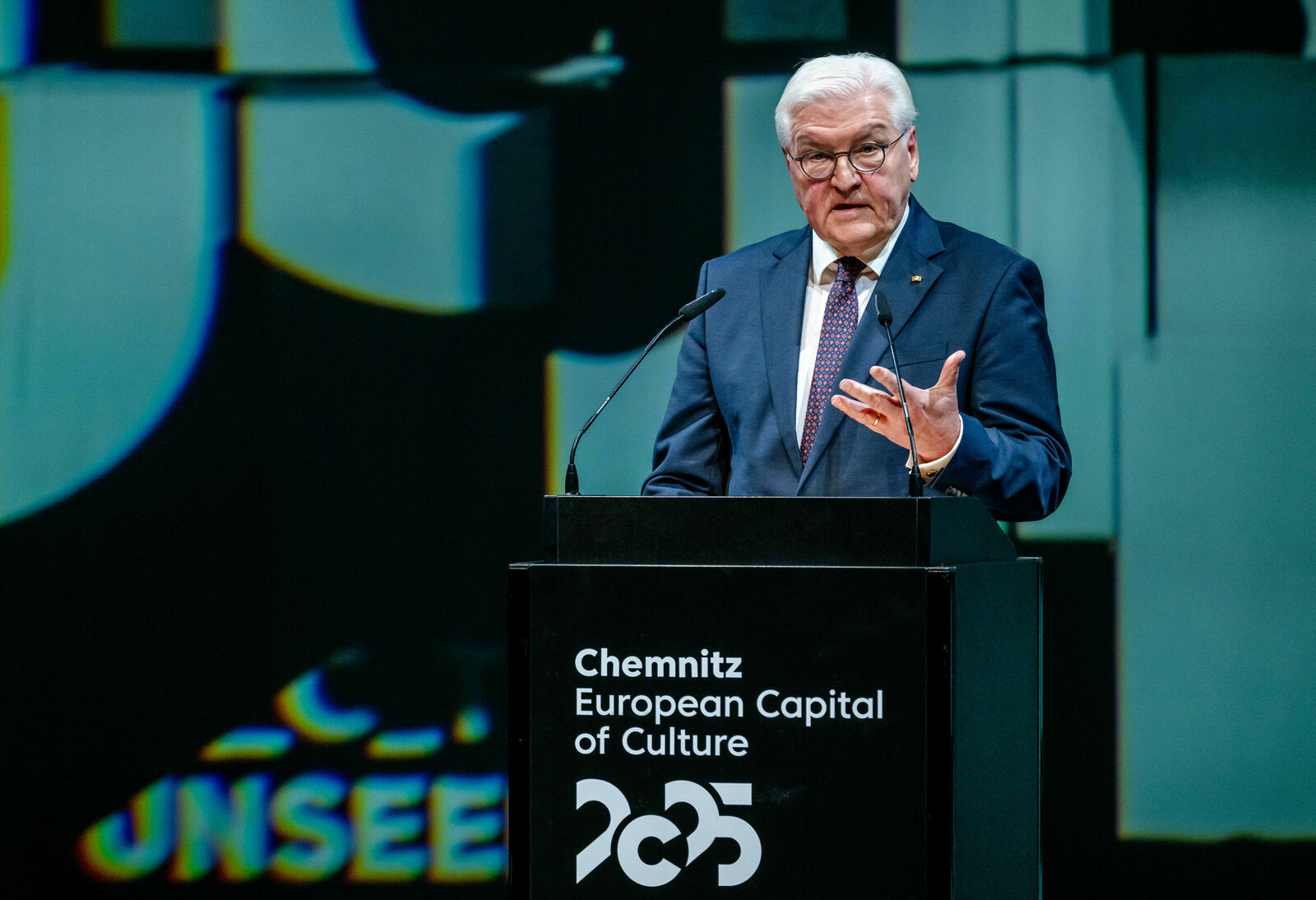 Der Bundespräsident Frank-Walter Steinmeier steht an einem Rednerpult und spricht während einer Veranstaltung zur Ernennung von Chemnitz zur Europäischen Kulturhauptstadt 2025. Auf dem Pult steht die Aufschrift "Chemnitz European Capital of Culture 2025" und im Hintergrund sind verschwommene, grafische Projektionen zu sehen.