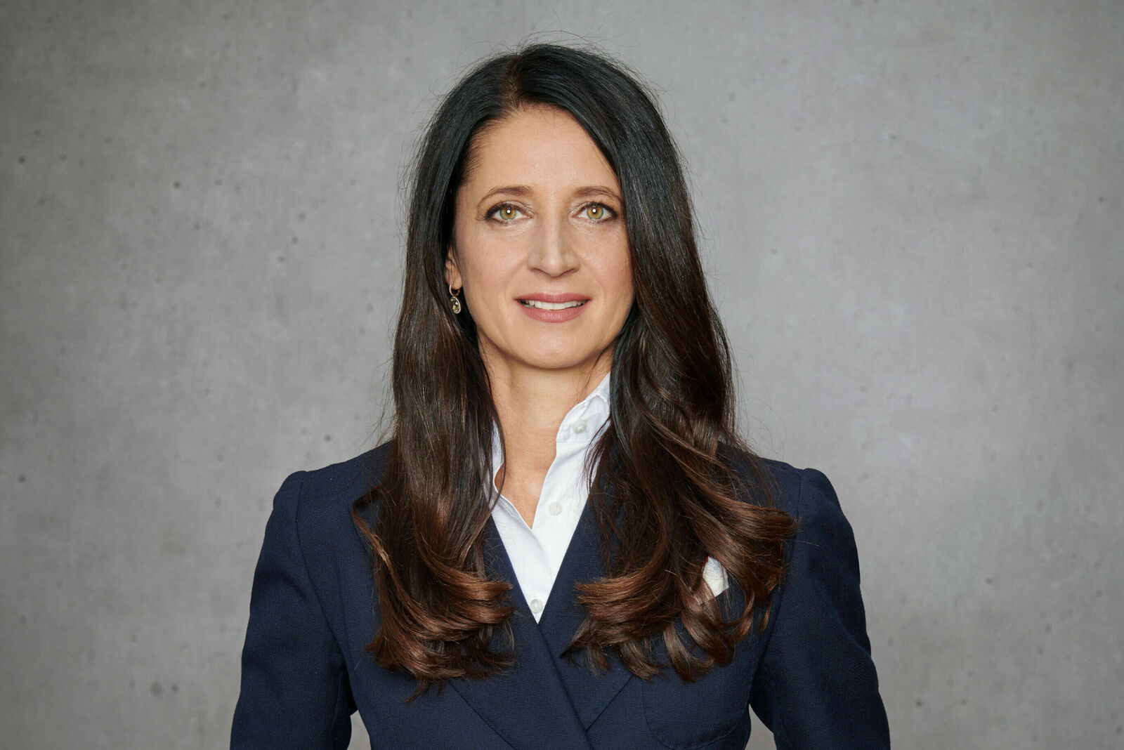 The image shows a professional woman with long dark hair, standing in a dark blue suit and white shirt in front of a gray wall. She has a friendly expression on her face.