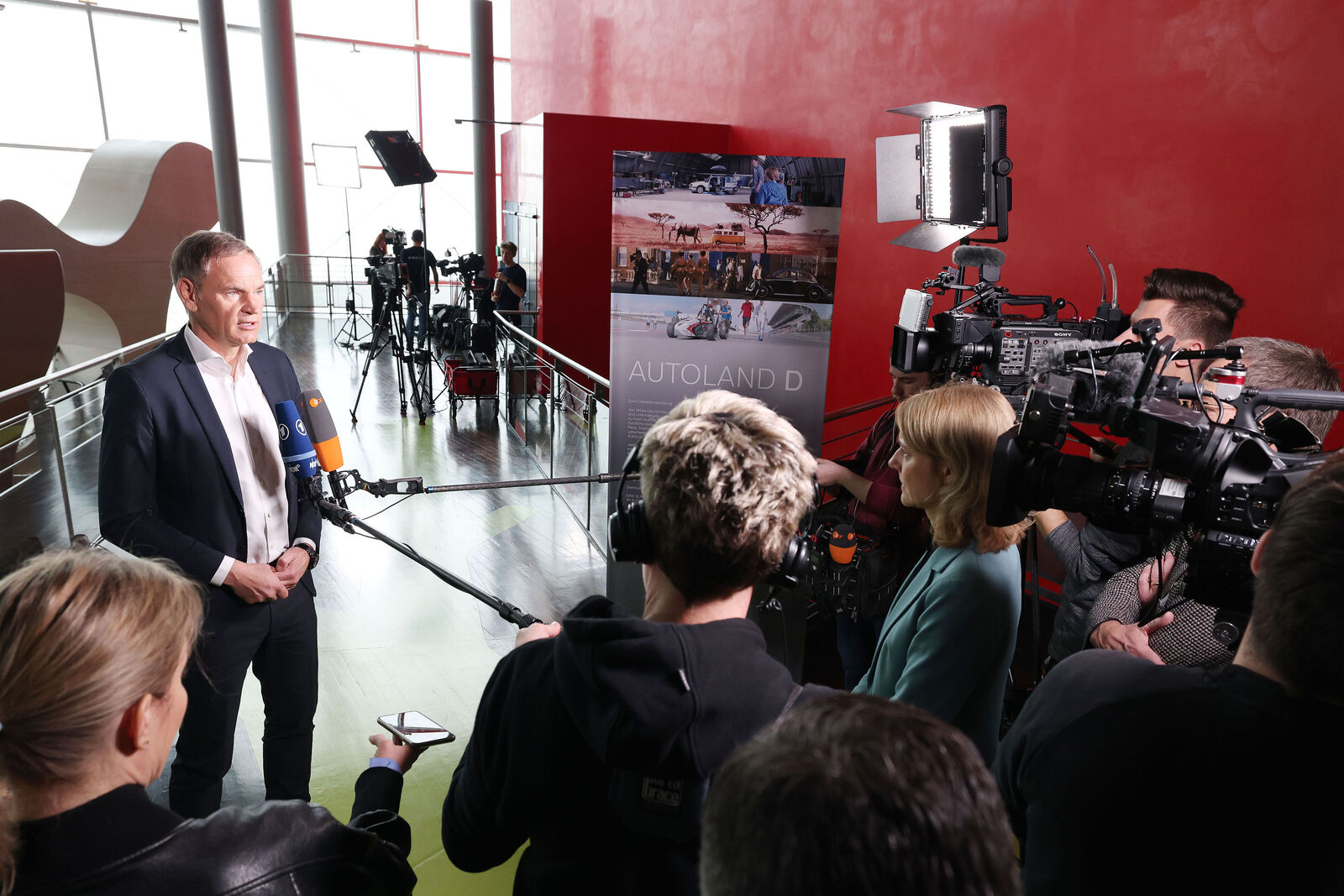 A man in a suit gives an interview in front of a group of journalists and cameramen. Microphones, cameras, and lighting equipment are directed at him. In the background, a poster with the title “AUTOLAND D” stands against a red wall.