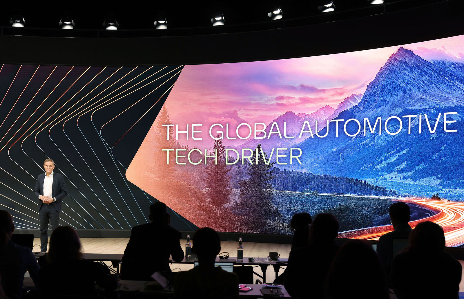 Ein Mann in Anzug präsentiert auf einer Bühne vor einem großen, gebogenen Bildschirm. Die Leinwand zeigt ein Landschaftsbild mit Bergen und einer kurvigen Straße mit Lichtspuren, begleitet vom Text „THE GLOBAL AUTOMOTIVE TECH DRIVER“. Im Vordergrund sitzt das Publikum.