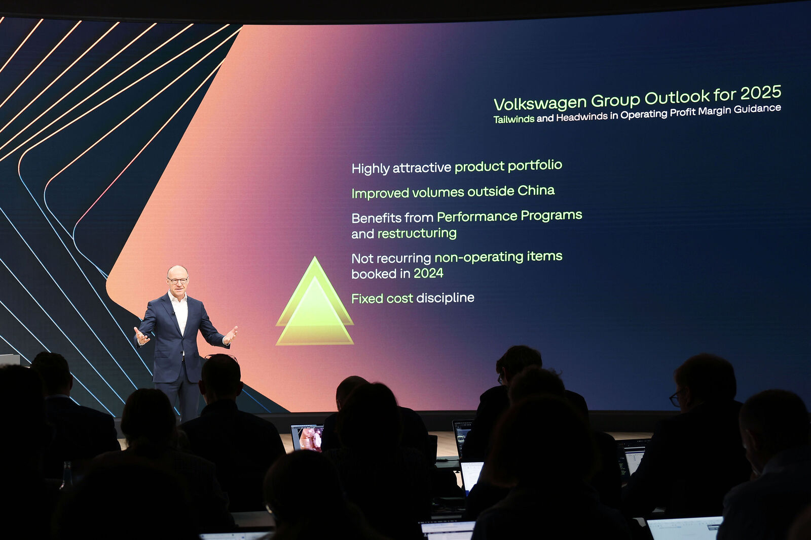 A man in a suit presents on a stage in front of a large screen. The slide displays the “Volkswagen Group Outlook for 2025” with several strategic points in green and yellow text, along with a stylized pyramid. The audience is seated in the foreground, using laptops.