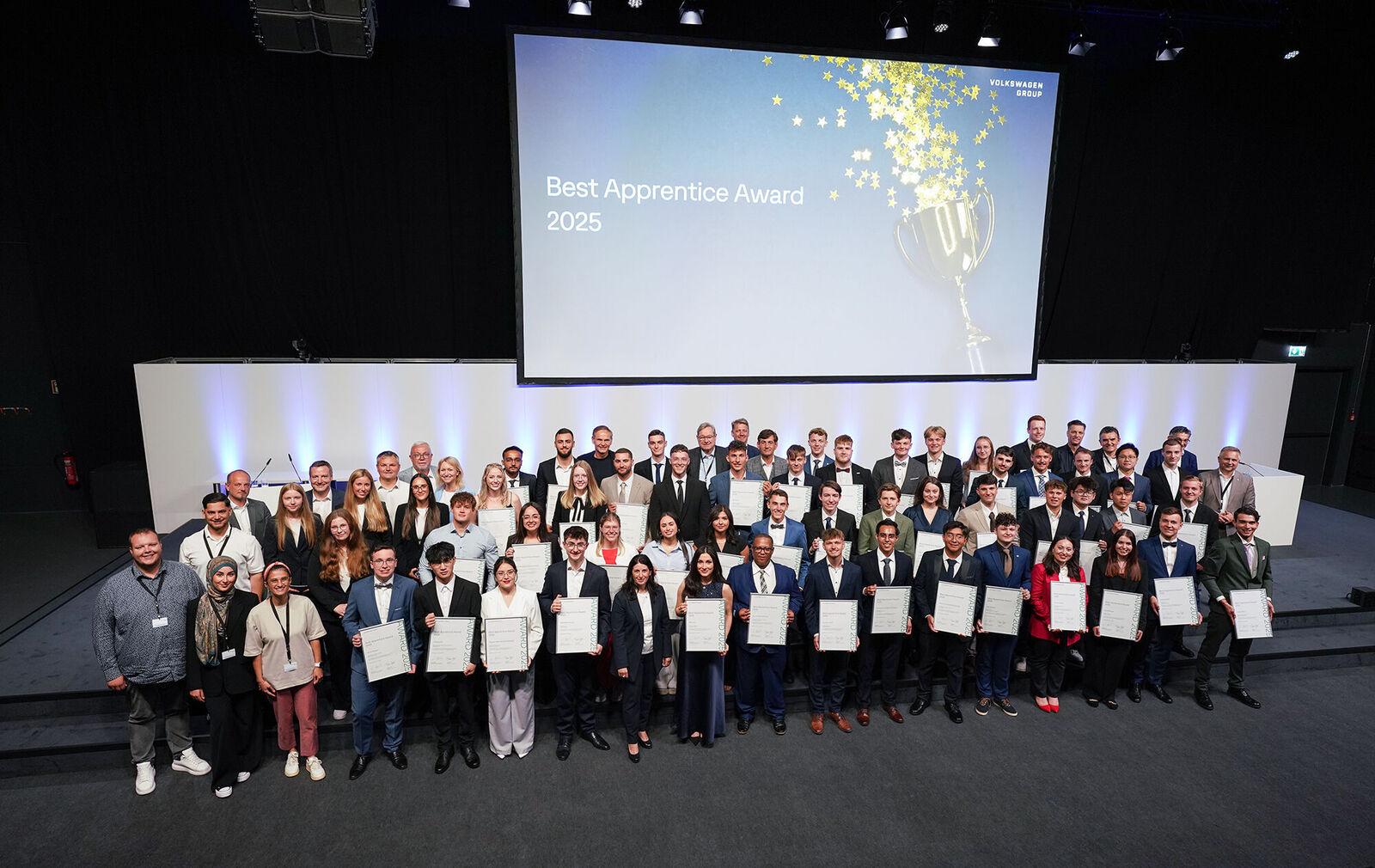 Gruppenfoto der Gewinnerinnen und Gewinner des „Best Apprentice Award 2025“ der Volkswagen Group. Rund 60 junge Menschen stehen auf einer Bühne, halten ihre Urkunden in den Händen und lächeln in die Kamera. Im Hintergrund zeigt eine große Leinwand den Titel der Veranstaltung.