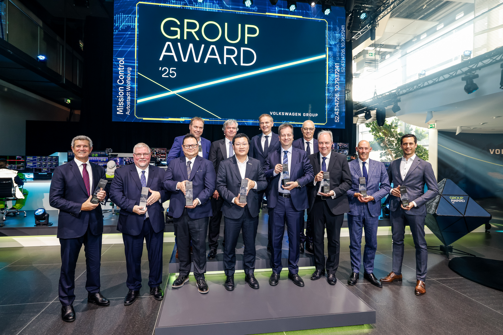 Gruppe von elf Männern in Anzügen mit Trophäen in der Hand bei der Volkswagen Group Award-Verleihung 2025. Im Hintergrund große Leinwand mit der Aufschrift „GROUP AWARD '25“ und „Volkswagen Group“.