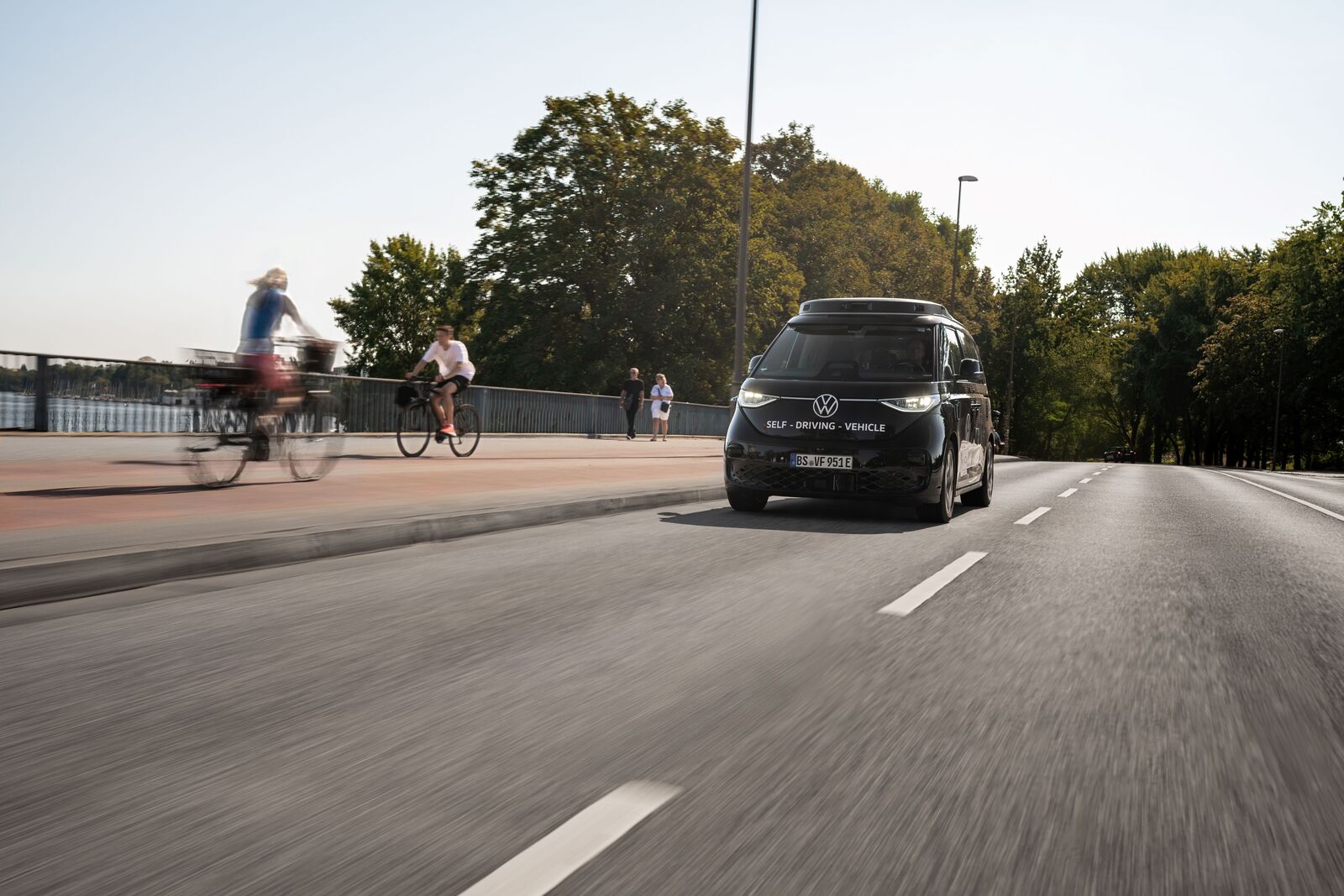 Selbstfahrendes VW-Fahrzeug fährt auf einer städtischen Straße, während Radfahrer und Passanten am Rand unterwegs sind.
