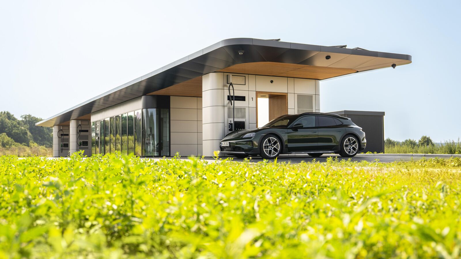 Ein schwarzes Elektroauto steht vor einer modernen, futuristisch gestalteten Ladestation mit flachem Dach und großen Glasflächen. Im Vordergrund ist eine grüne Wiese zu sehen, im Hintergrund blauer Himmel.