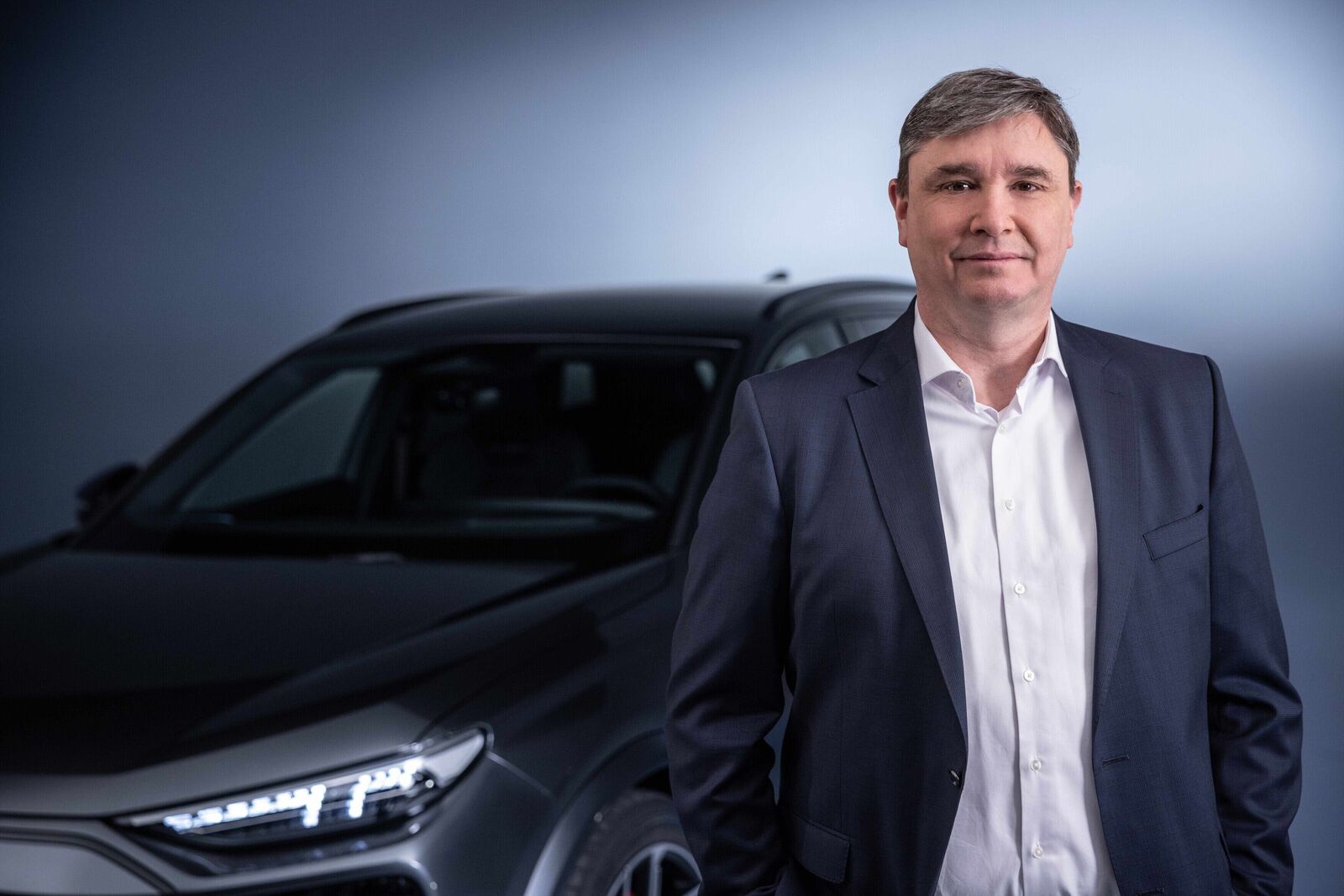 A man in a dark suit and white shirt stands in front of a black car, smiling at the camera.