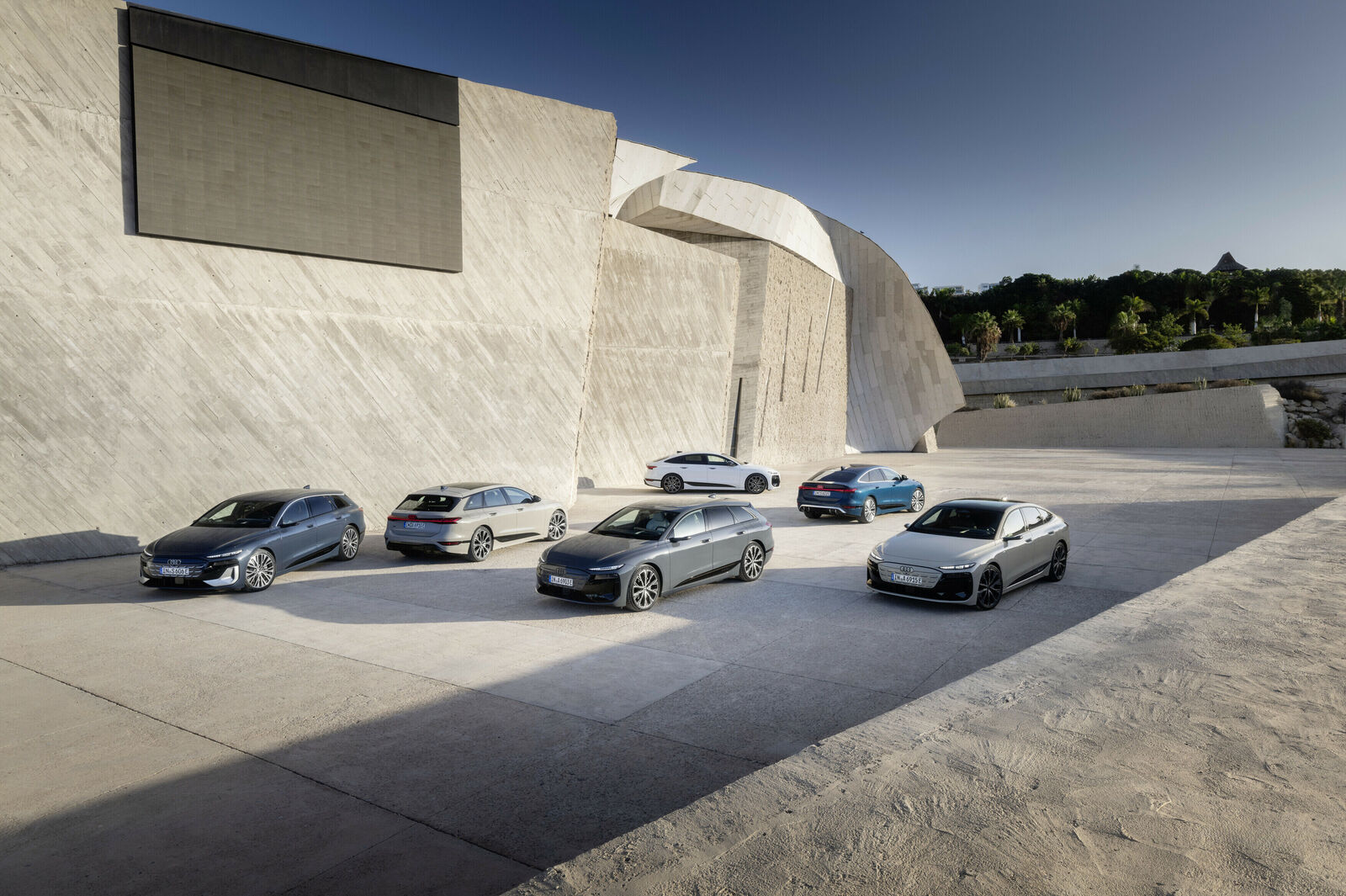 Six modern Audi vehicles are arranged in a geometric formation on a spacious plaza in front of a monumental, futuristic concrete building.