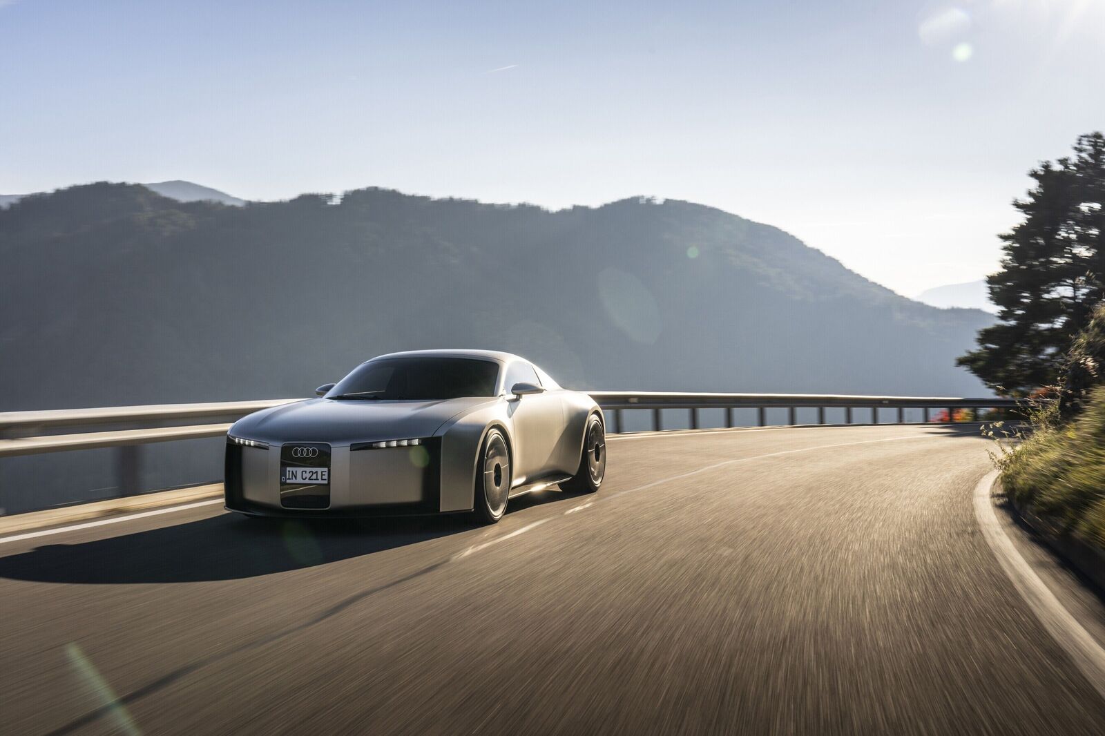 A futuristic silver Audi sports car drives along a winding mountain road in bright sunlight, with hills and mountains visible in the background.