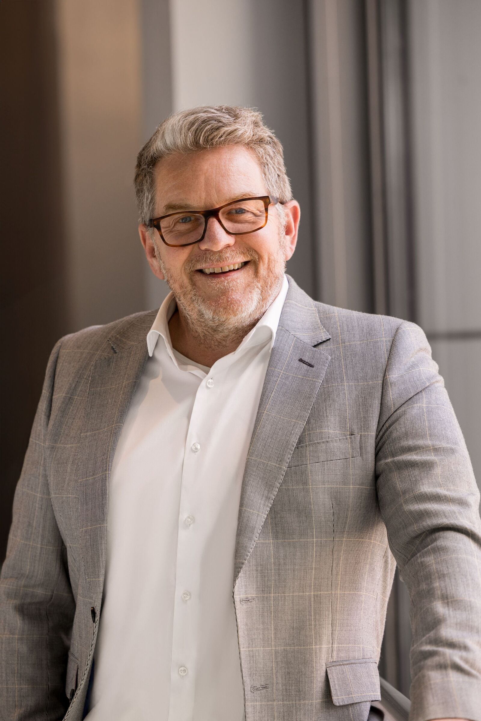 Portrait of a smiling man with glasses, gray blazer, and white shirt by a window.