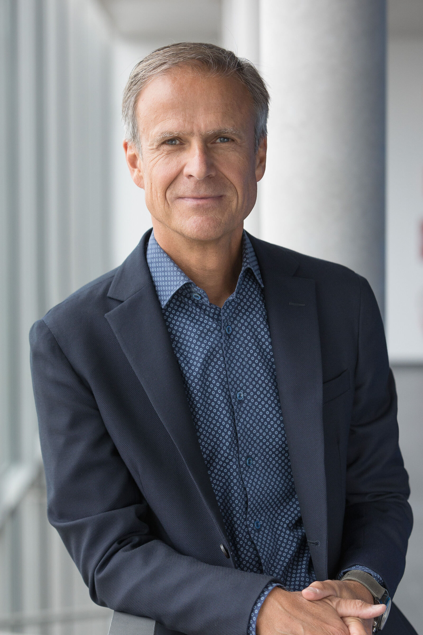 Portrait of a man in a navy blazer and patterned shirt, relaxed by a railing in a bright office setting.