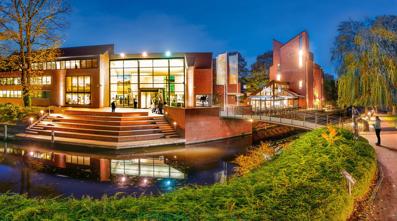 Abendliche Außenansicht eines beleuchteten Backstein-Museums am Wasser mit Treppen, Brücke und Café; einige Besucher gehen vorbei.