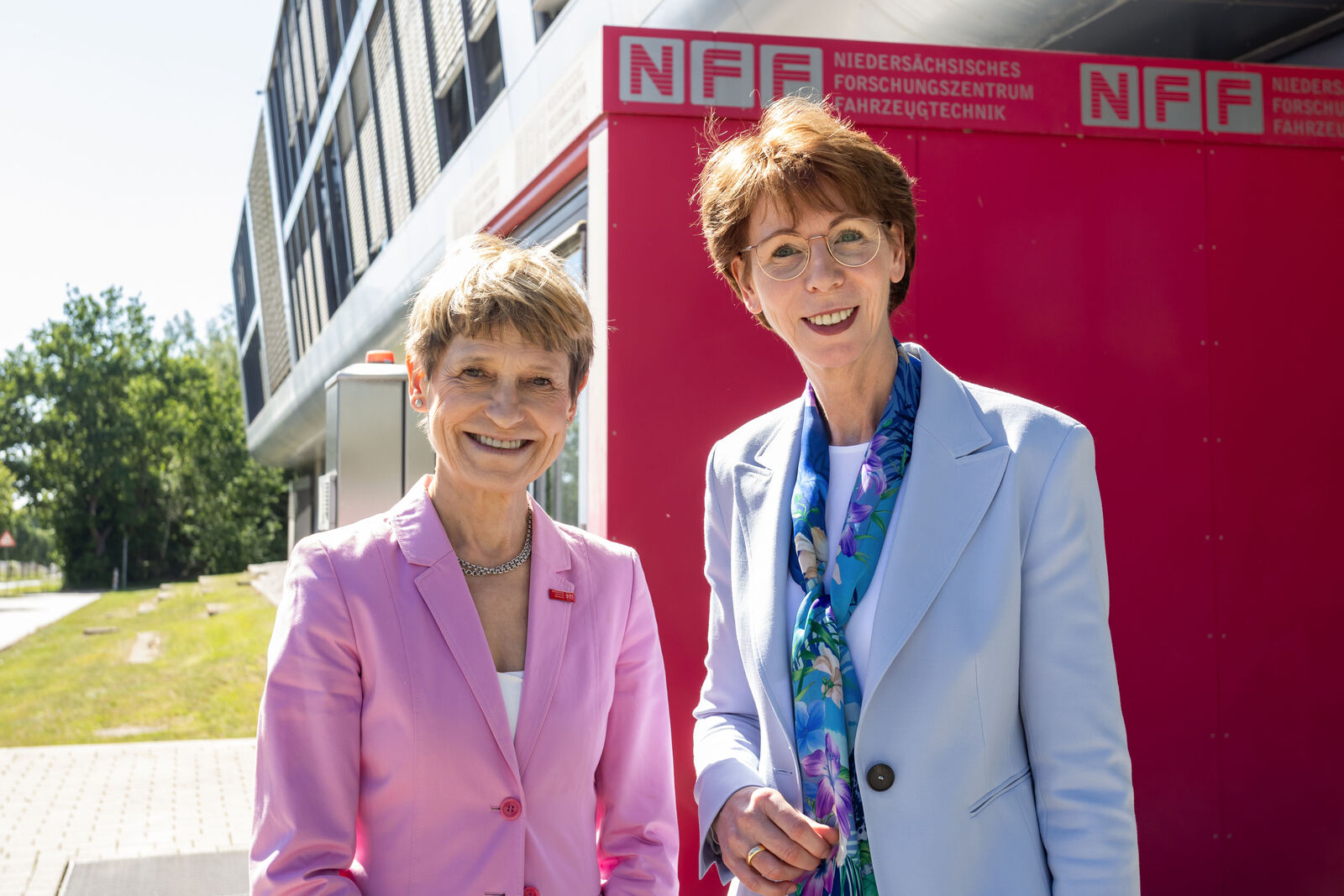 Zwei Frauen in Businesskleidung stehen lächelnd vor einem Gebäude mit NFF-Logo.