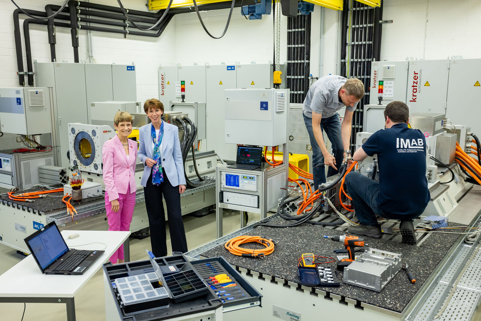 Zwei Frauen in einem technischen Labor, während Techniker an Maschinen und Kabeln arbeiten.