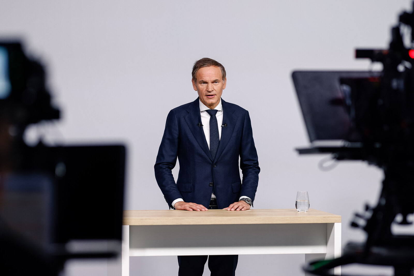 Oliver Blume im blauen Anzug am hellen Tisch; im Vordergrund unscharfe Kameras der TV-Produktion zur VW Hauptversammlung.