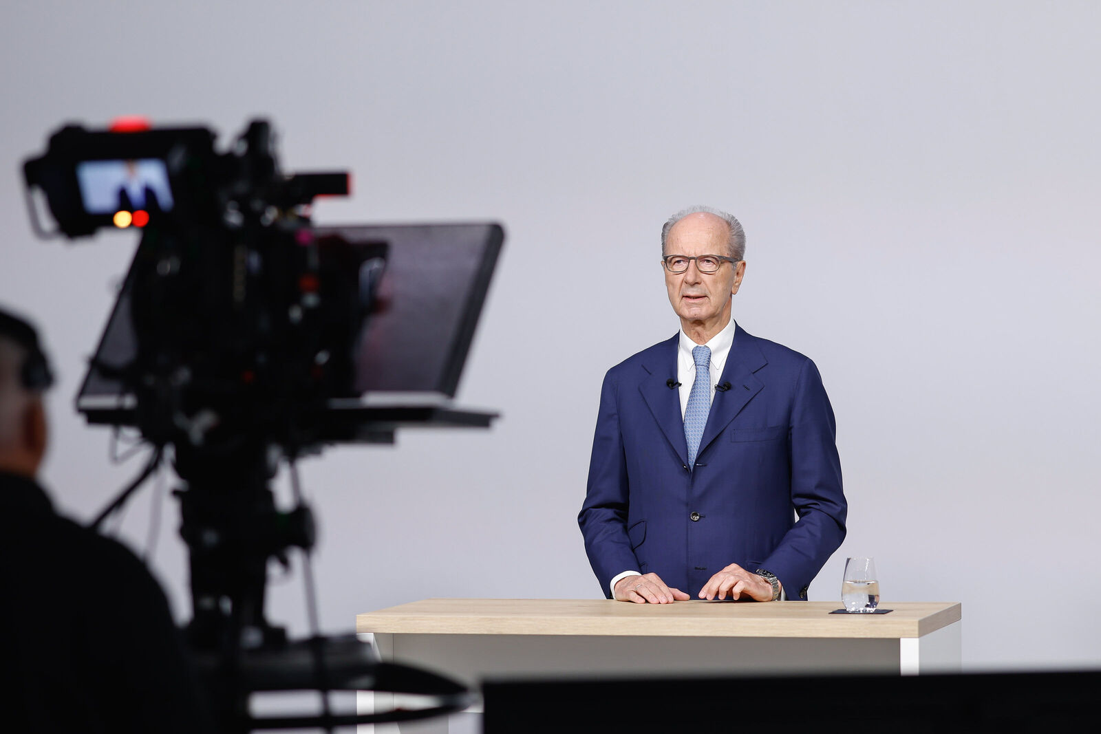 Hans Dieter Pötsch in a studio speaking behind a table to a camera; filming equipment in the foreground, a glass of water on the table.