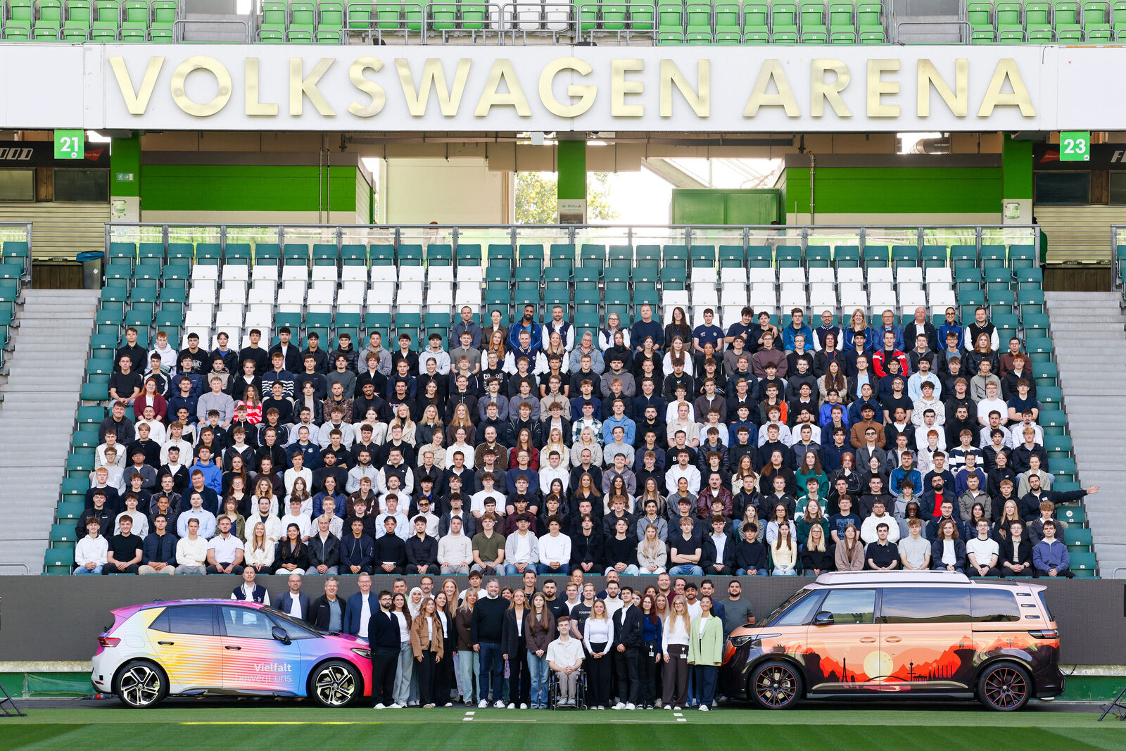 Großes Gruppenfoto in der Volkswagen Arena: Mehrere Personen sind auf den Stadiontribünen versammelt. Vor ihnen stehen auf dem Spielfeld zwei Volkswagen Fahrzeuge mit farbenfroher Folierung. Über der Gruppe ist der Schriftzug ‚Volkswagen Arena‘ sichtbar.