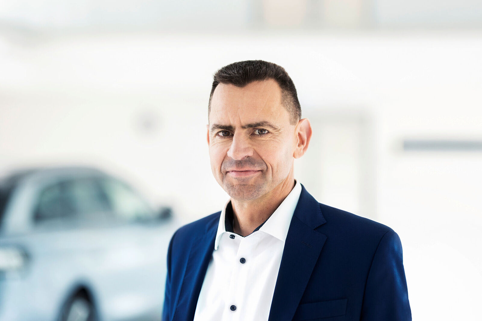 Businessporträt von Martin Sander in blauem Anzug und weißem Hemd in einer hellen, modernen Innenraumumgebung. Im unscharfen Hintergrund ist ein Fahrzeug zu erkennen.