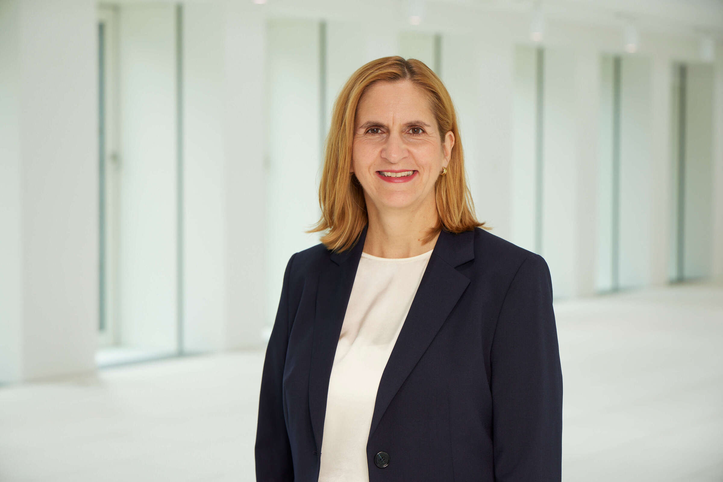 Portrait of Erika Rasch wearing a dark blue blazer and a light-colored top, standing in a bright, modern office corridor with large windows in the background.