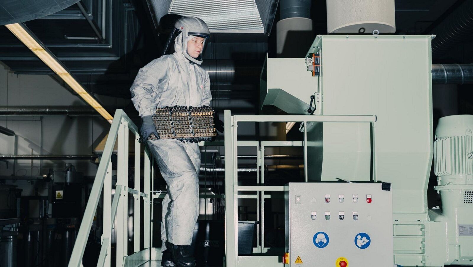 Arbeiter im Schutzanzug trägt Batteriezellen zu einer Recyclinganlage in einer Industriehalle.