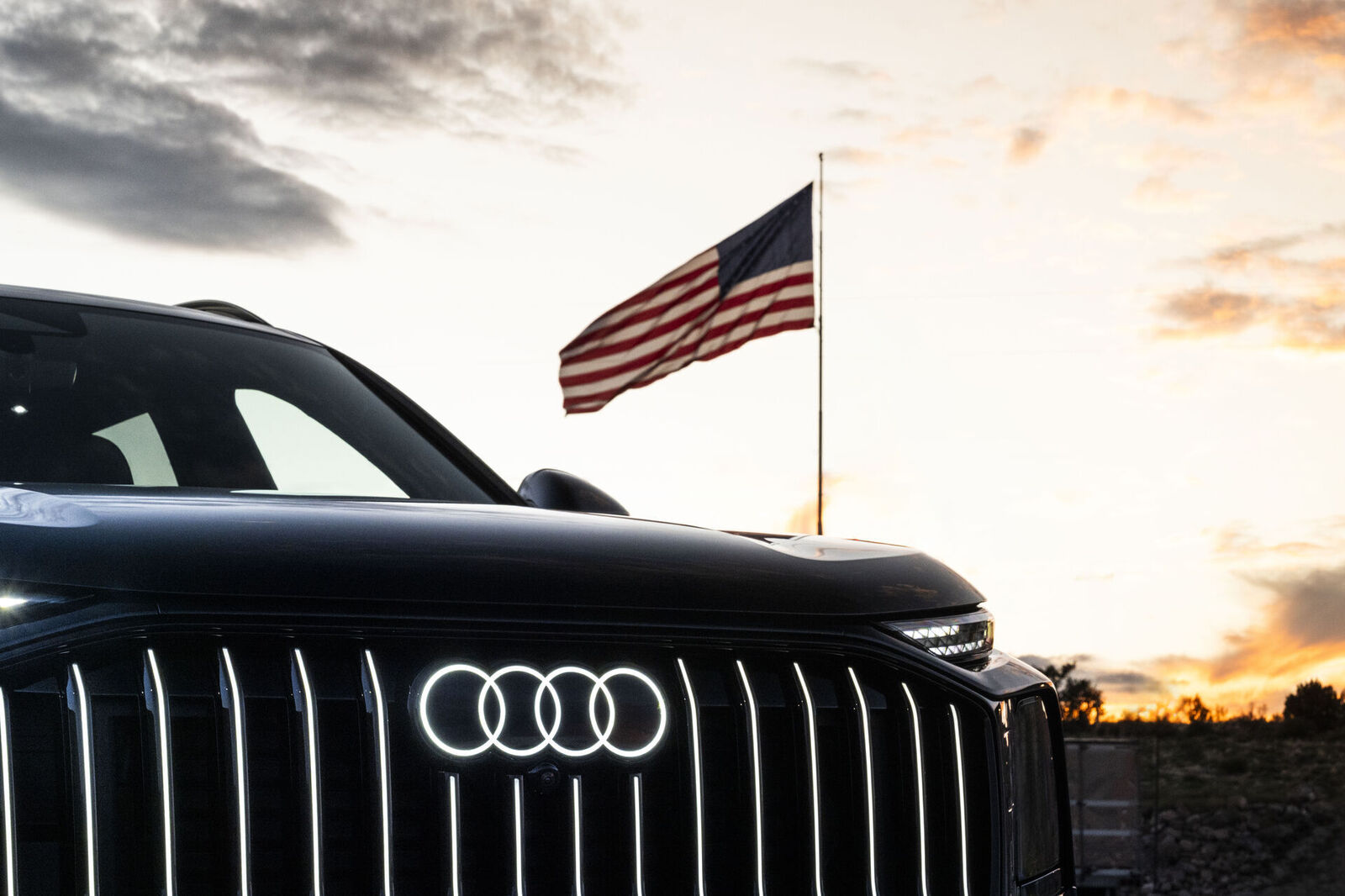 Front eines schwarzen Audi mit beleuchtetem Logo vor einer amerikanischen Flagge bei Sonnenuntergang.